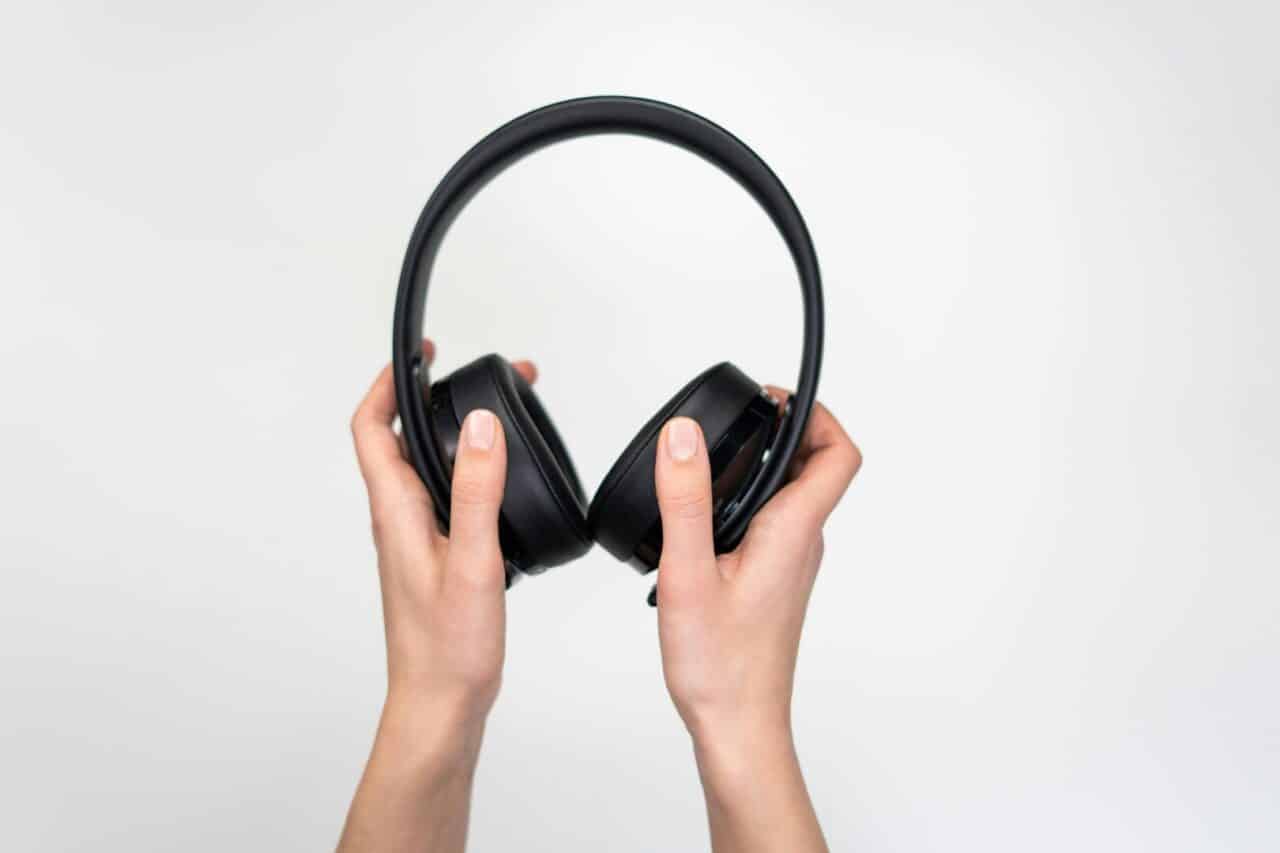 A pair of over-ear headphones held up by a pair of hands in front of a pale background.