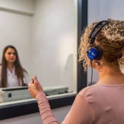 Woman holding button to indicate hearing a sound during a hearing test with audiologist behind partition across from her