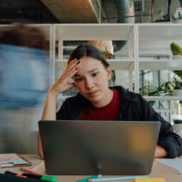 Young woman feeling overwhelmed and stressed while working intently on a laptop in a bustling office environment filled with activity